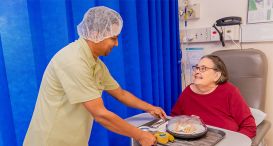 Nurse serving a meal to a smiling patient in hospital bed.